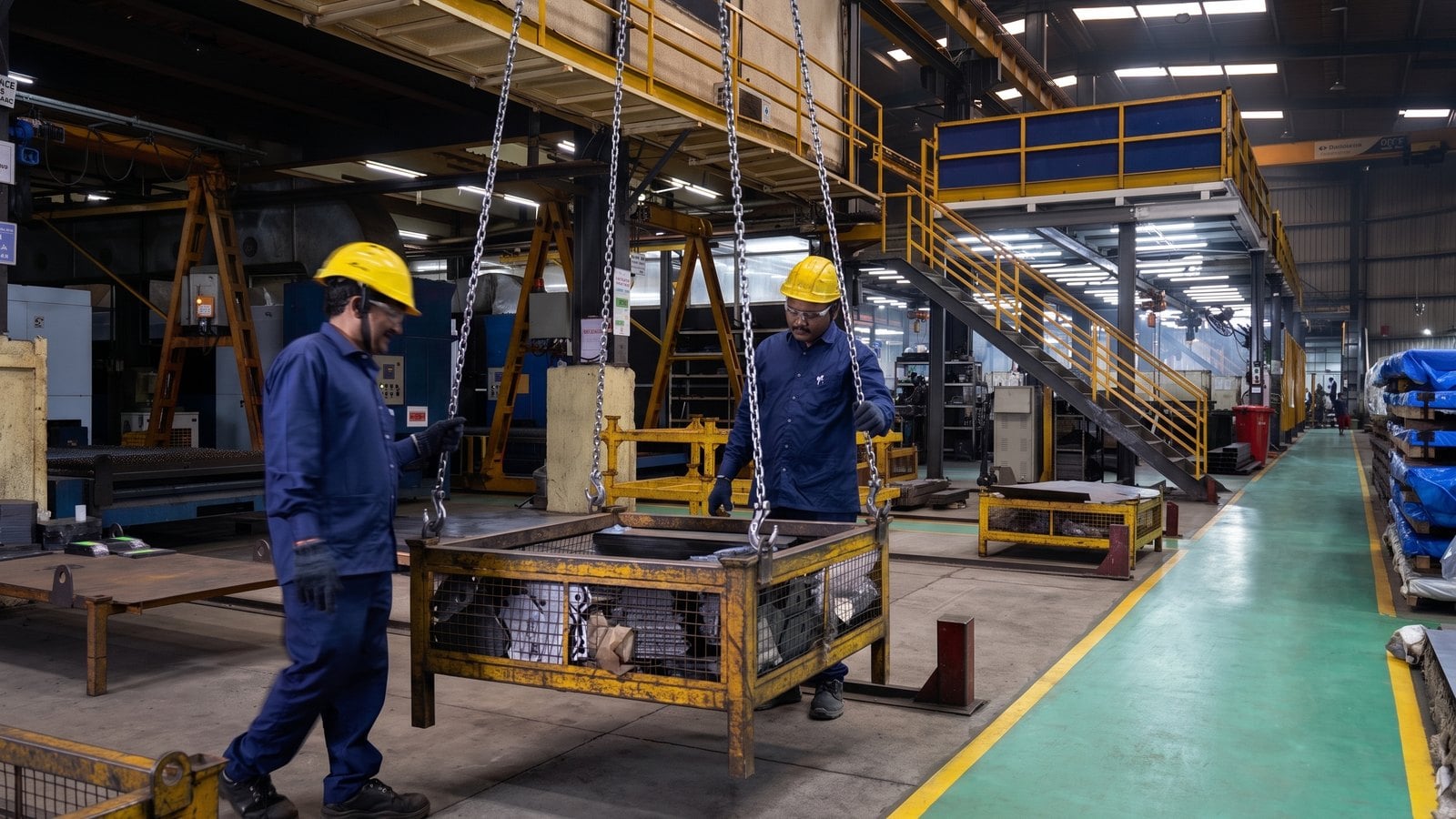 Two workers guiding a crane-lifted stillage of parts down the plant aisle